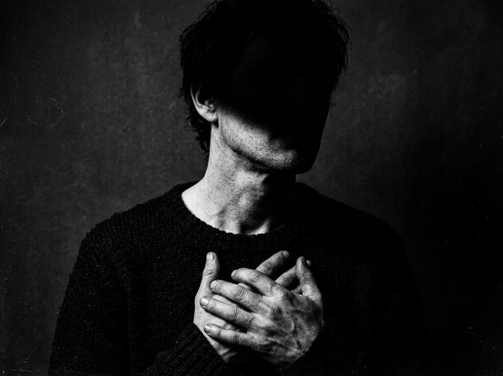 Dark portrait of a man with his face hidden in shadow and hands over his chest, representing detachment from validation, inner focus, obsession, discipline, and emotional control.