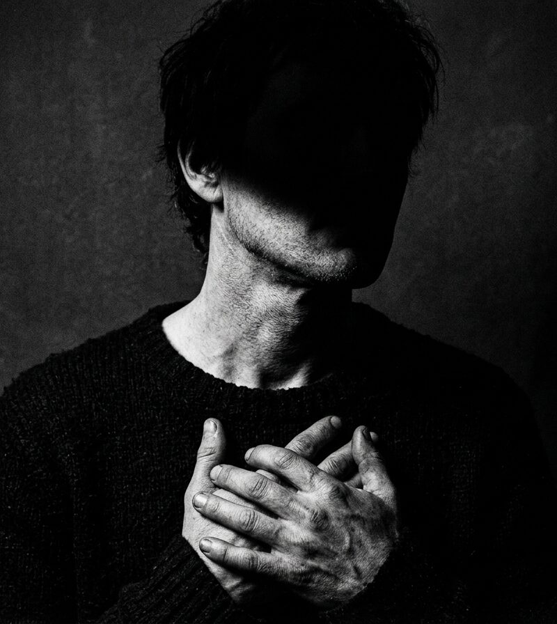 Dark portrait of a man with his face hidden in shadow and hands over his chest, representing detachment from validation, inner focus, obsession, discipline, and emotional control.